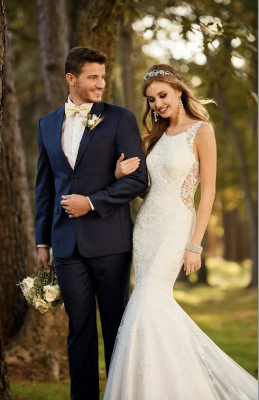 Image shows a male model wearing a navy suit smiling at a female model wearing a sleeveless mermaid wedding gown
