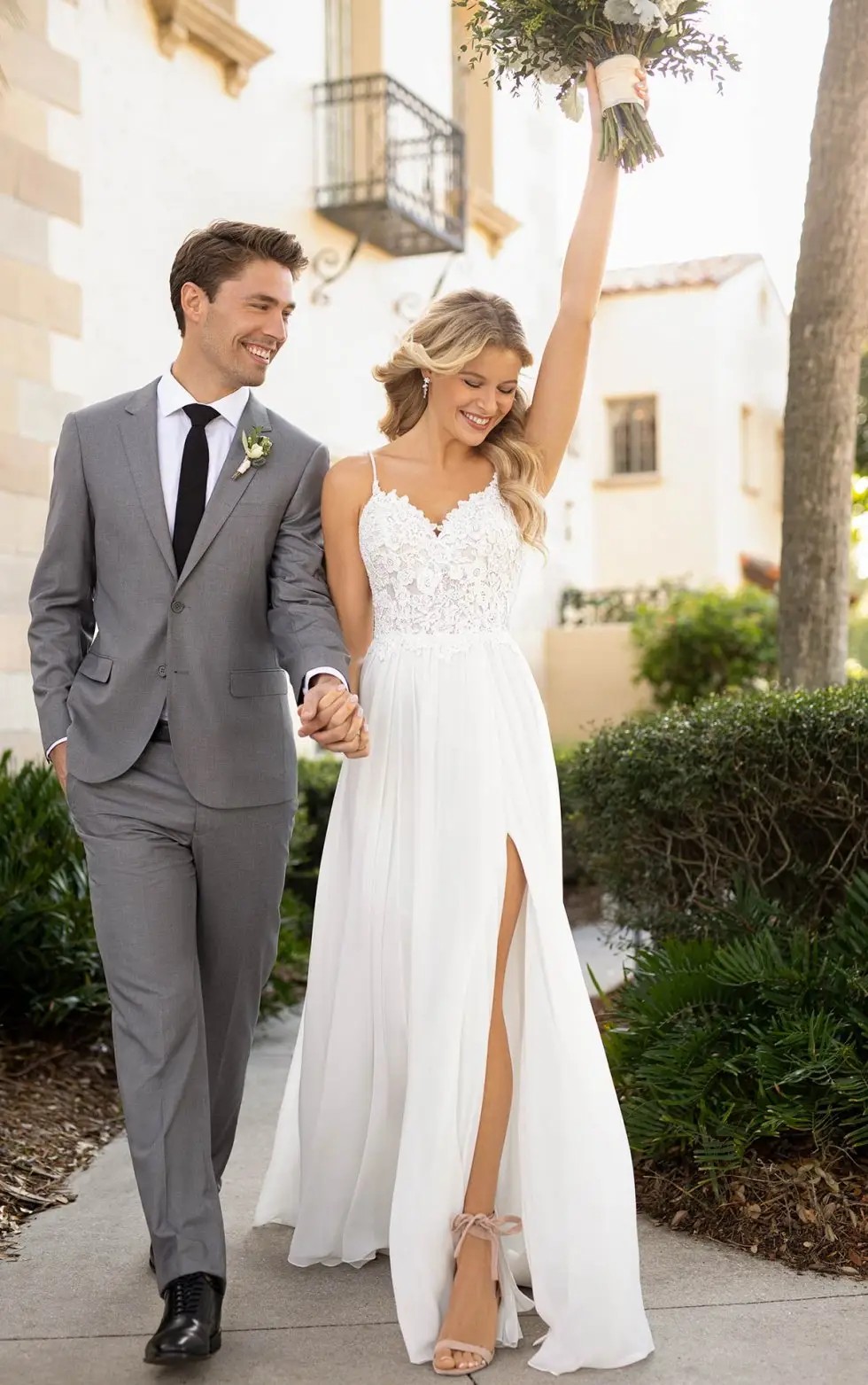 Image shows a male model in a grey suit smiling and holding hands with a female model wearing a sleeveless a-line wedding dress with spaghetti straps and a high slit