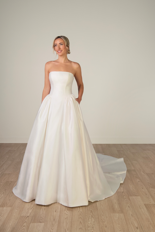 A woman is wearing a strapless white wedding dress and smiling, standing on a wooden floor in a neutral-colored room.