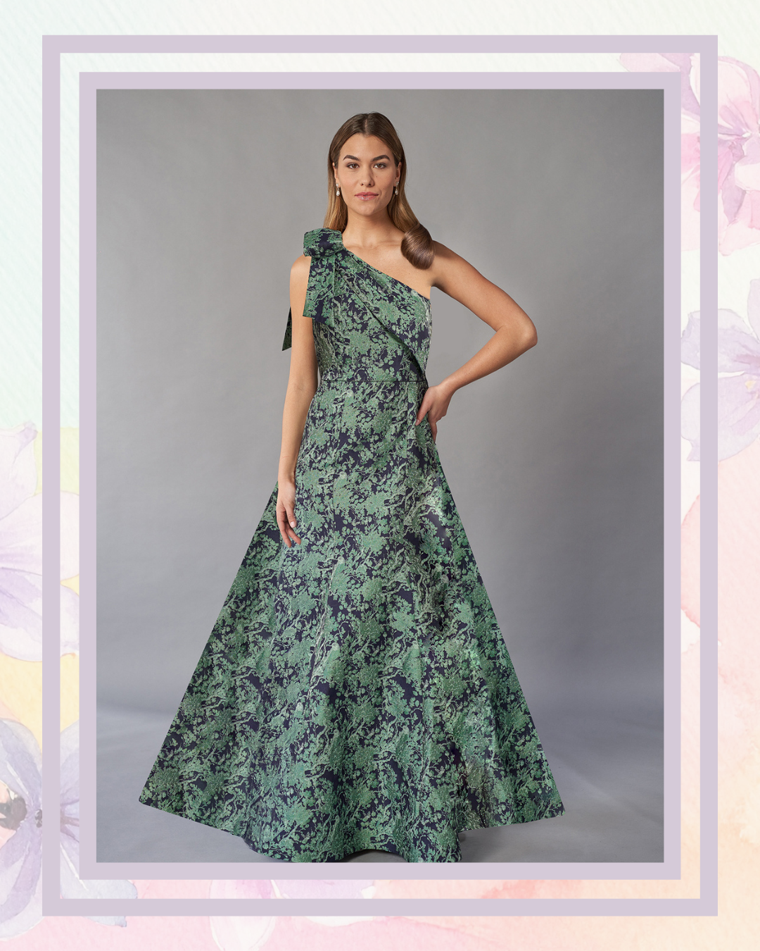 A model is wearing a one-shoulder, floor-length dress featuring a floral pattern in shades of green and blue. The dress has a fitted bodice and flared skirt, with a bow detail on one shoulder.&nbsp;