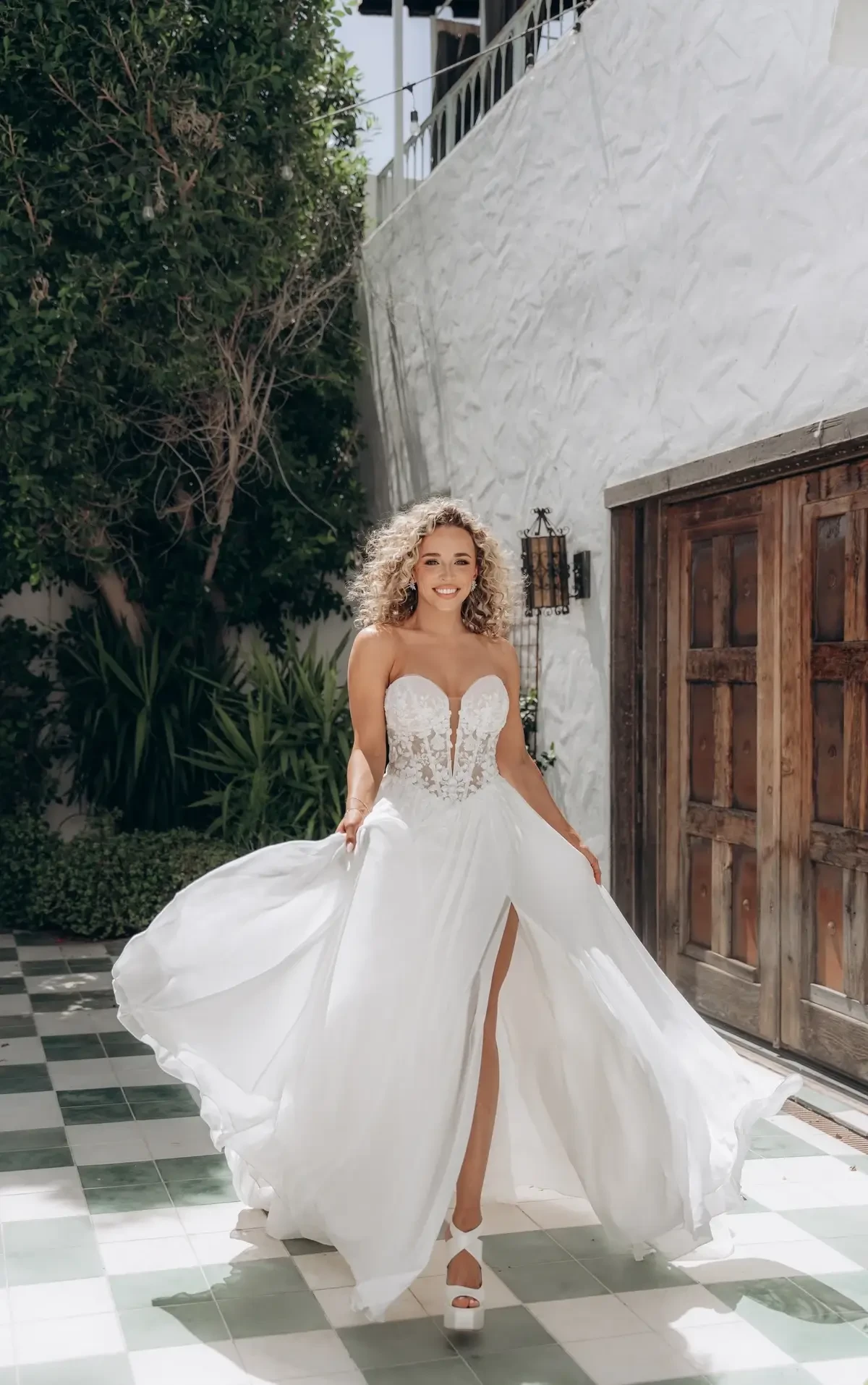 A woman in a white strapless wedding dress with a high slit is smiling as she walks on a checkered floor, surrounded by greenery and a wooden door.
