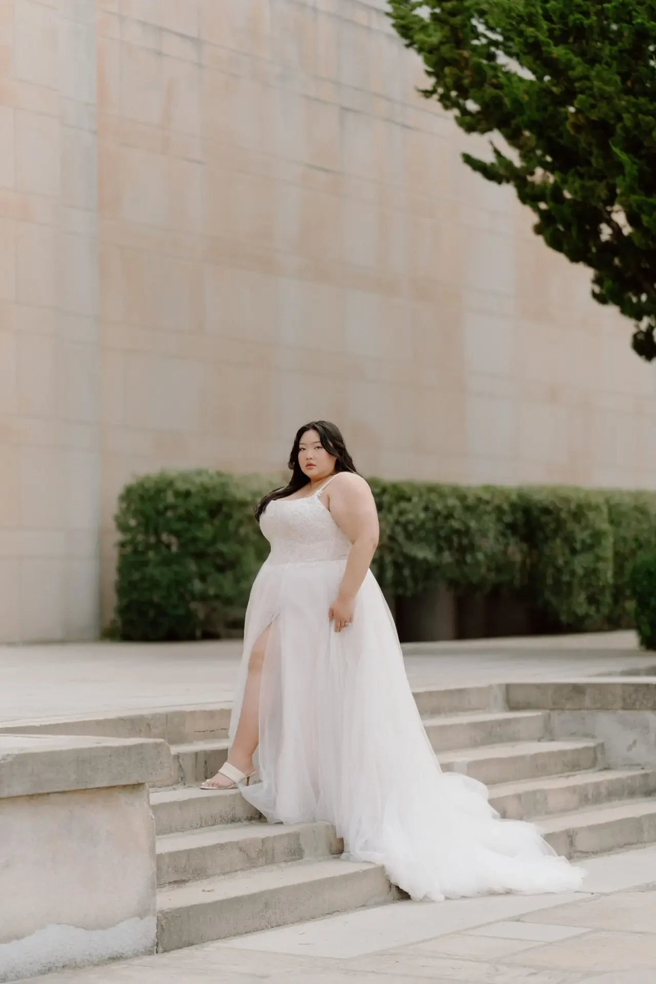 A woman wearing a white dress with a thigh-high slit stands on stone steps in front of a simple, textured wall and greenery.