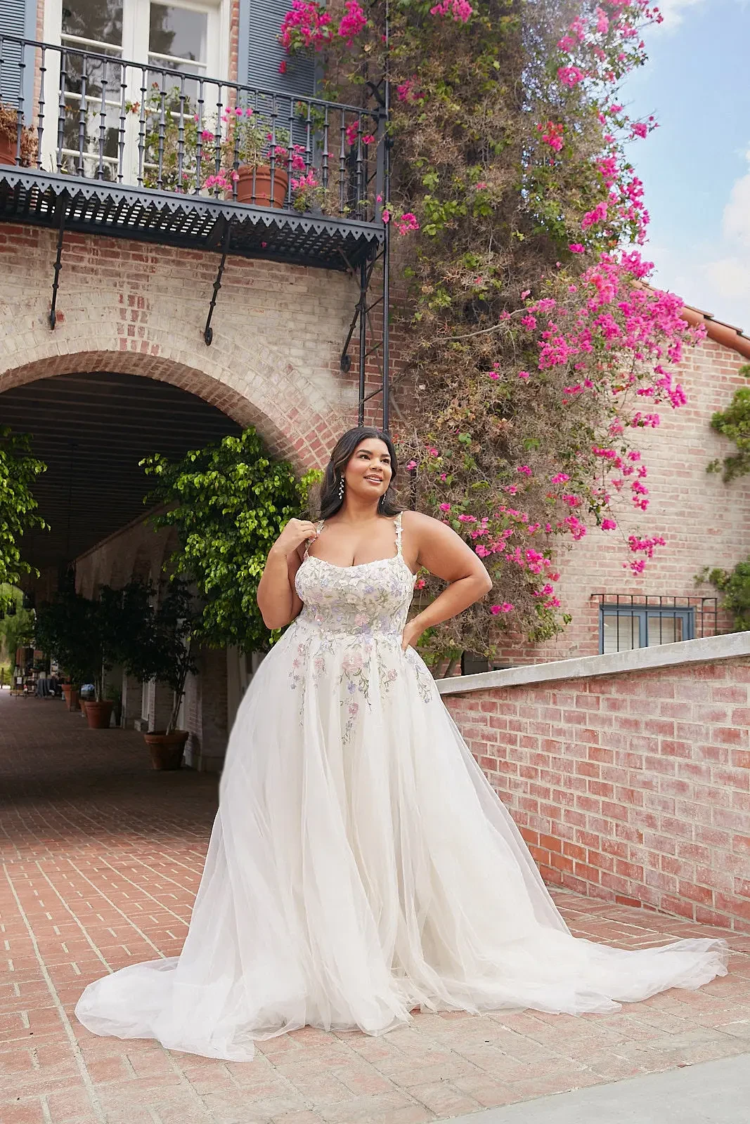 A woman in a flowing white gown stands elegantly in a picturesque outdoor setting, framed by blooming pink flowers and brick arches.