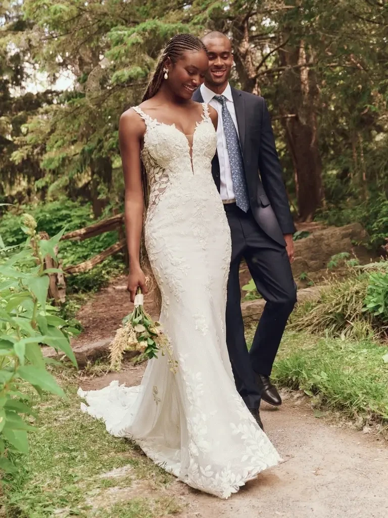 A couple walking through a forested area. The woman is wearing a fitted, lace wedding dress and holding a small bouquet, while the man is dressed in a suit.