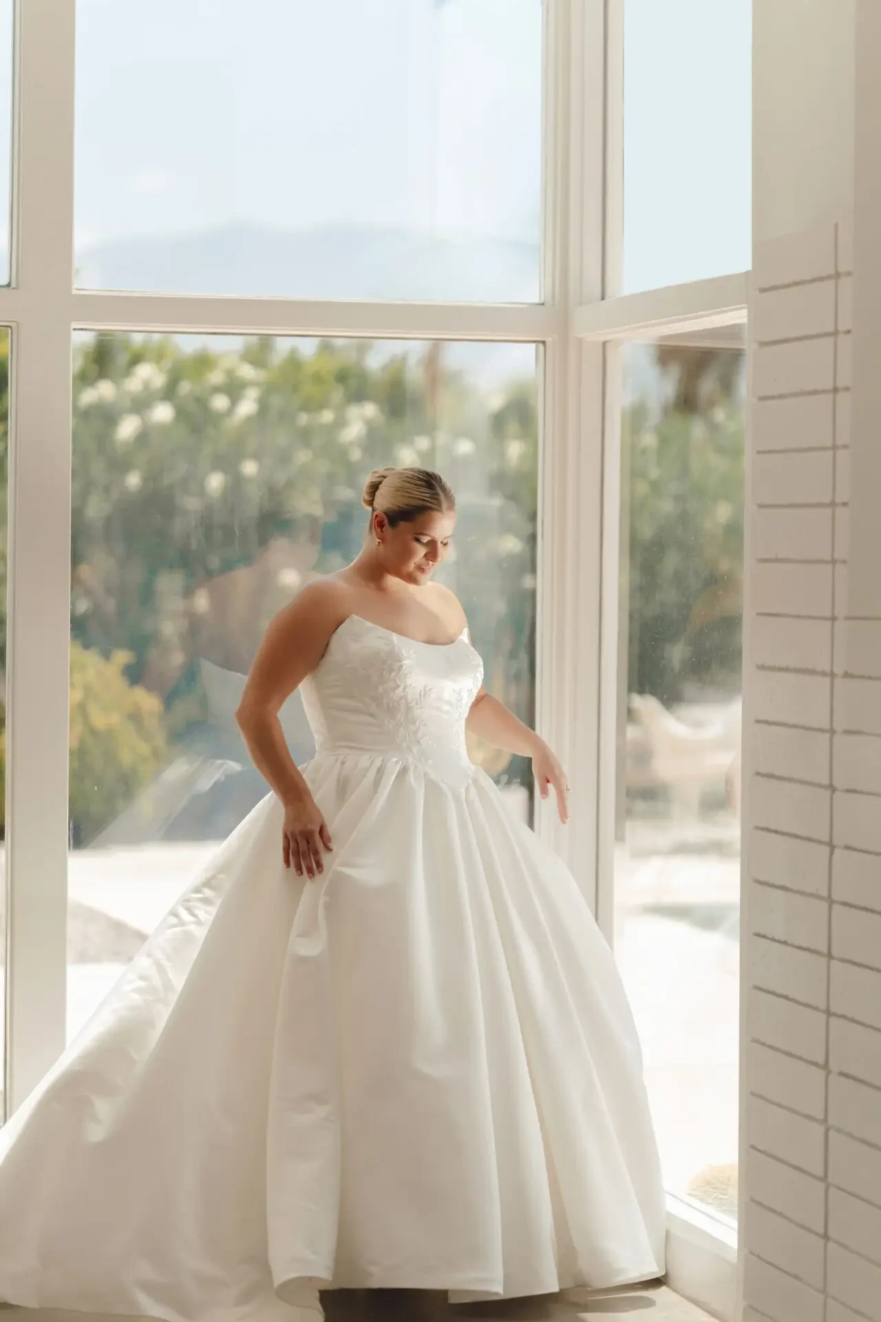 A woman in a wedding dress stands near a large window, with soft natural light illuminating the scene.