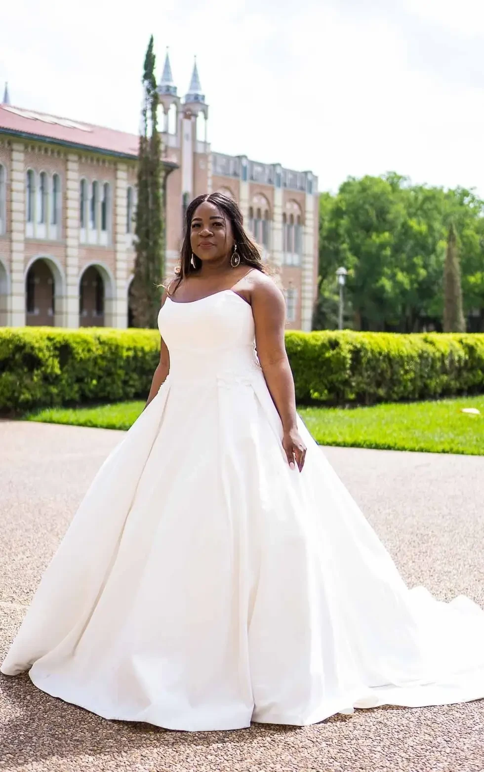 A woman in a white wedding dress standing on a path with greenery and a building in the background.