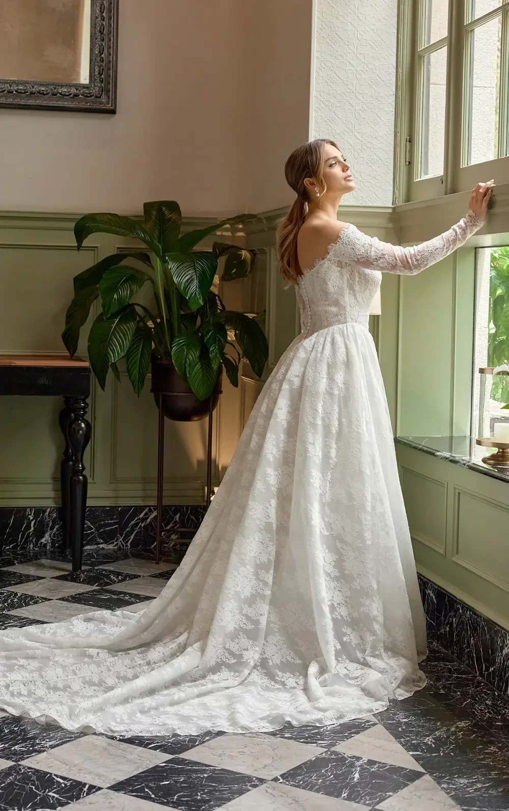A woman in an elegant off-shoulder white wedding dress standing in a stylish interior with a chandelier overhead.