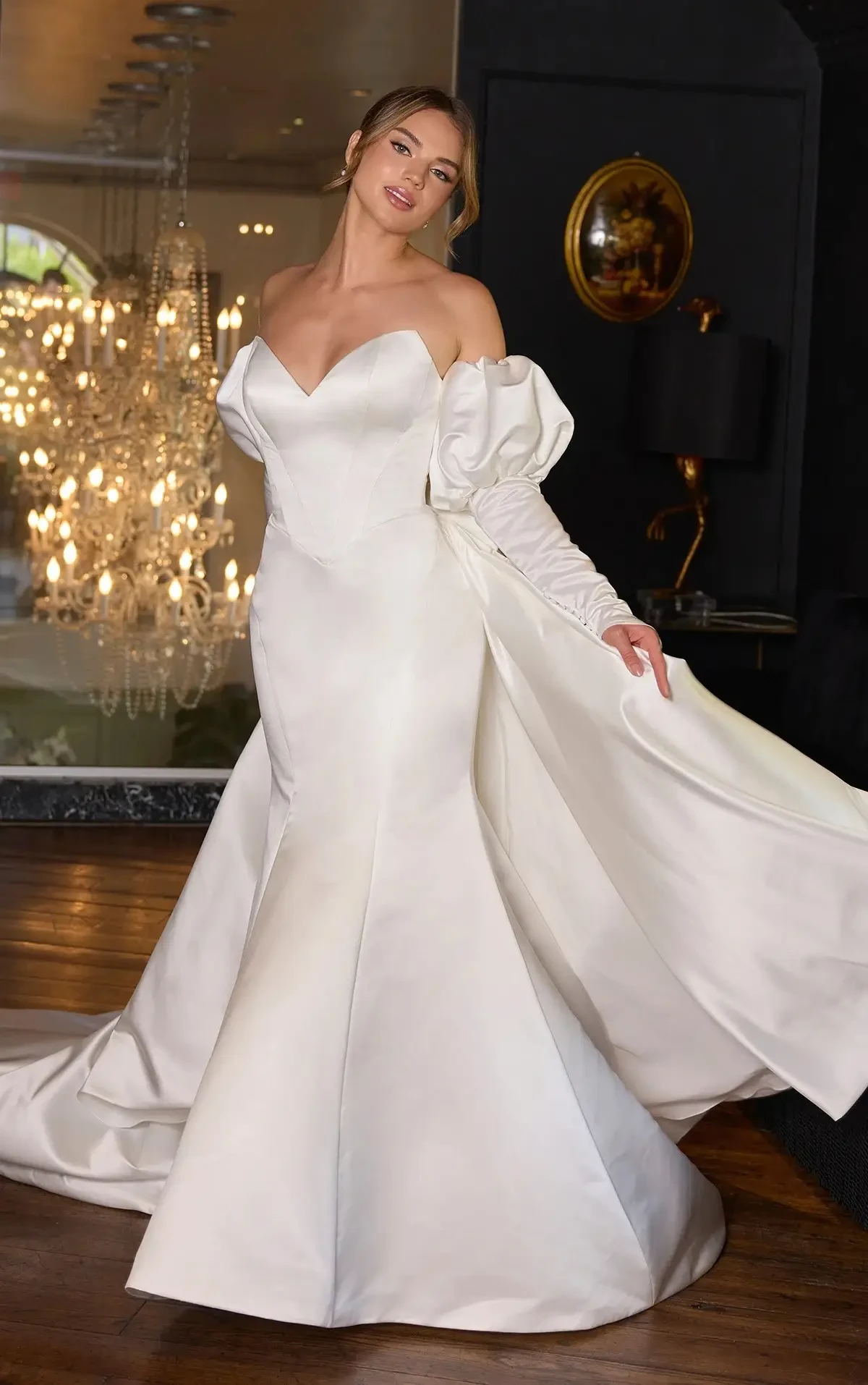 Woman in an elegant white off-shoulder wedding gown with voluminous sleeves stands in a dimly lit room, accented by a sparkling chandelier.