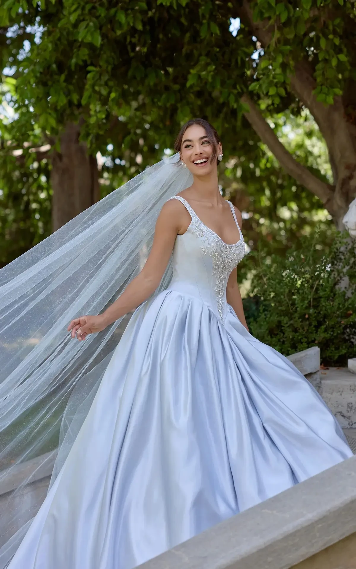 Bride in a flowing white gown and long veil smiles joyfully outdoors, surrounded by lush green trees and soft natural light, conveying happiness and elegance.